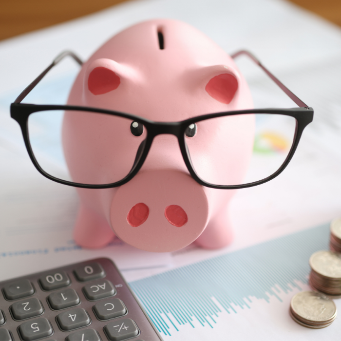 a piggy bank wearing glasses sits next to a calculator, some stacked coins, and spreadsheets